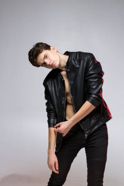 Deri ceketli yakışıklı genç adam stüdyoda duvarın arkasında poz veriyor. Çıplak bir gövdede siyah elbiseli genç bir eşcinsel portresi. İnce Uzun Kafkas Erkek Modeli.