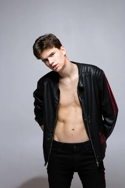 Handsome Athletic Male Model Wearing Leather Jacket on Naked Torso. Sexy young guy with a beautiful body in a biker jacket on a red background in the studio. Fashionable young Man in leather coat.