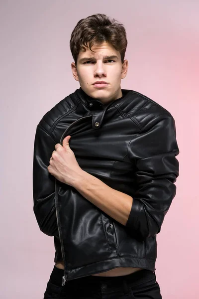Fashionable Handsome young man in leather jacket posing in the studio against the background of the wall. Portrait of a young gay man in black clothes on a naked torso. Thin tall Caucasian Male Model.