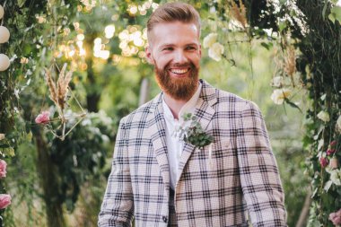 Beyaz tenli, şık sakallı damadın portresi. Eski moda takım elbiseli, güneşli havada açık havada poz veriyor. Ekose ceketli, sakallı yakışıklı bir adam. Düğünde kırsal tarzda bir erkek model. Kareli ceketli damat..