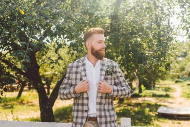 Klasik kıyafetler içindeki yakışıklı sakallı adam. Parktaki düğün gününde ekose ceketli şık damadın portresi. Köylü hipster portresi. Sakallı adam. Kareli ceketli şık saç kesimi olan bir model.