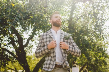 Kızıl sakallı bir adamın düğün portresi ve modaya uygun giyinmiş hippi saç stili. Sakallı damat bahçede ekose ceketli poz veriyor. Doğada düğün gününde erkek kırsal stil..