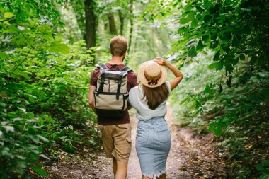 Ormanda el ele yürüyen bir çift, arka manzara. Macera, seyahat, turizm, yürüyüş, insanlar. Ağaçların arasında yürürken sırt çantası taşıyan iki kişinin arka görüntüsü. Adam kız arkadaşını gezintide elleriyle tutuyor..