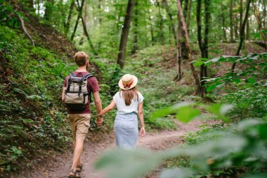 Ormanda el ele yürüyen bir çift, arka manzara. Macera, seyahat, turizm, yürüyüş, insanlar. Ağaçların arasında yürürken sırt çantası taşıyan iki kişinin arka görüntüsü. Adam kız arkadaşını gezintide elleriyle tutuyor..