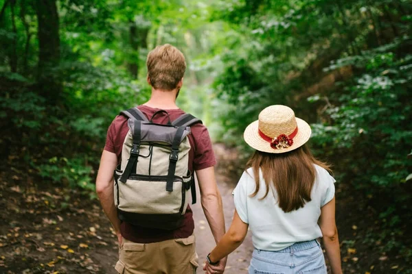 Guy and girl walk together in wood along the trail, holding hands. Back view. Happy couple holding hands and walking forest path. Hikers walk in wooded area with a backpack in summer.