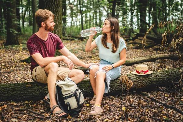 森の散歩中に休憩を取るハイカー 若い白人男性と女性は倒木の上に座り プラスチック製の透明なボトルから水を飲む 二人の旅行者が森の中で水を飲む — ストック写真