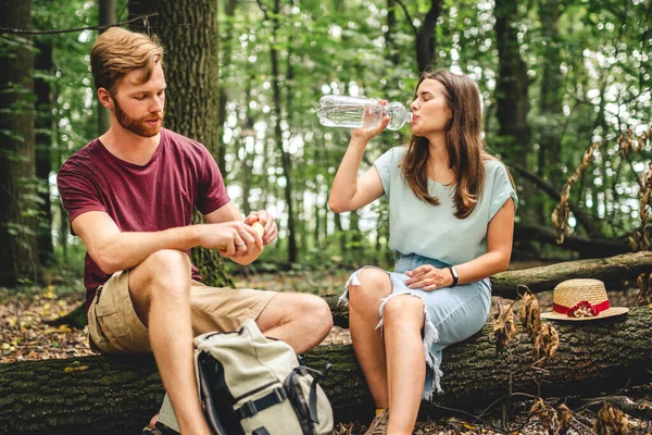 Caucasian couple stopped to rest while walking in the forest, young people sit on a fallen tree trunk drinking water from a bottle. Active rest, hike in a wooded area. Thirst and water balance.