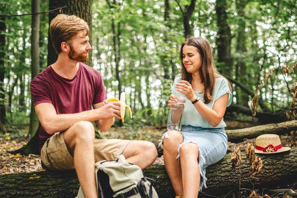 young couple sat to rest during forest walk along path to fallen tree, people drink water and eat banana fruit. Tourists break for snacks and replenishment water balance. Food and drinks in nature.