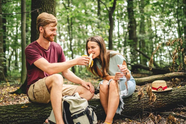 People eat banana and drink water from plastic bottle while sitting on log in wood. Couple hikers take break for food and drink in forest on fallen tree trunk. Stop for picnic, trail forest walk.