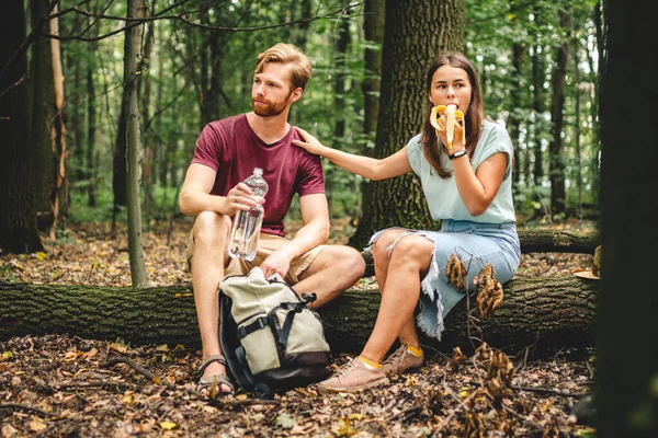 木に座っている間にバナナを食べ ペットボトルから水を飲む カップルのハイカーは 倒木の幹で食べ物や飲み物を休憩します ピクニック トレイルフォレストウォークのために停止 — ストック写真