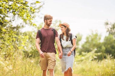 Kafkasyalı hippi çift kırsal yolda yürüyor. Sırt çantasıyla doğa parkında yürürken sevgili kızının elini tutan sakallı bir adam. Seyahat, yürüyüş, turizm ve insan konsepti.
