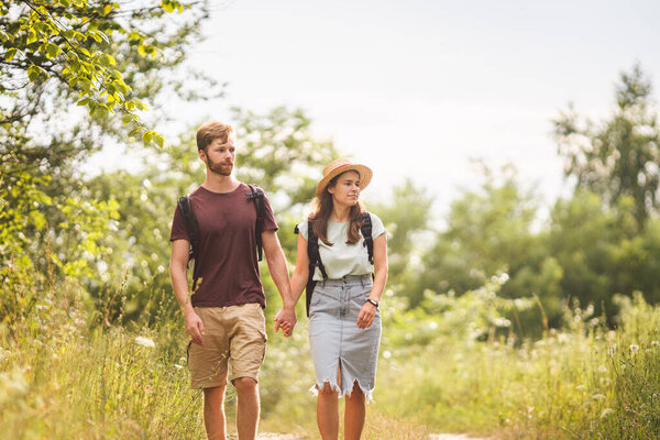 Caucasian hipster couple walking along path in countryside. man with beard holds the hand of his beloved girl while hiking in natural park with backpack. Travel, hiking, tourism and people concept.