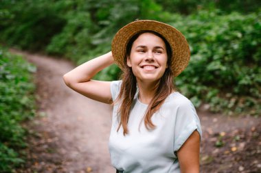 Güzel bir ormanda takılan heyecan verici bir kızın açık hava fotoğrafı. Eski hasır şapkalı neşeli genç bayan parkta poz veriyor. Ormanda yürüyen güzel genç bayan. Her adım bir maceradır..