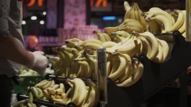 Üniformalı ve süpermarkette eldiven giyen bir işçinin yakın çekimi vitrindeki muzları ayırmaktır. Süpermarket çalışanı muz seçiyor. Markette organik olgun muz meyvesi