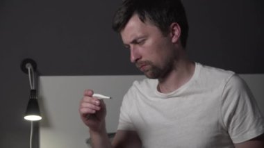 Evde grip olmuş genç bir adam var. Grip olmuş bir adam. Kötü durumda olan hastalıklı bir erkek. Hasta adam vücut ısısını termometreyle ölçüyor. Vücut ısısı ölçümü. Ateşim var mı?