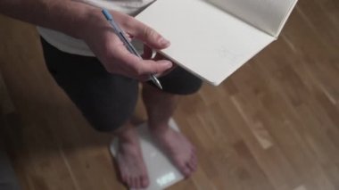 İnce adamın çıplak bacakları beyaz banyo terazisine basar. Erkek evde antrenmandan sonra kilosunu kontrol eder. Adam ağırlık ve yağ oranını dijital ağırlık ve sağlıklı yaşam tarzına göre kaydediyor.