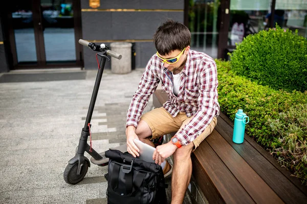 Man Putting Laptop in Backpack outside sitting near electric transport scooter. Caucasian man packing laptop in backpack. Male pulling laptop out backpack. Person with computer sitting on wooden bench