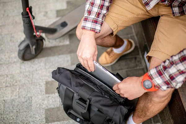 Man Putting Laptop in Backpack outside sitting near electric transport scooter. Caucasian man packing laptop in backpack. Male pulling laptop out backpack. Person with computer sitting on wooden bench