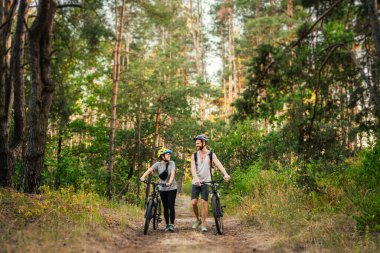 Bisiklet süren ve orman yolunda yürüyen neşeli bir çift. Bisikletli mutlu çift parkta yürüyor, konuşuyor ve gülüyor. Kafkasyalı genç adam ve kadın ormanda bisiklet sürüyor..
