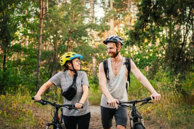 Bisiklet süren ve orman yolunda yürüyen neşeli bir çift. Bisikletli mutlu çift parkta yürüyor, konuşuyor ve gülüyor. Kafkasyalı genç adam ve kadın ormanda bisiklet sürüyor..