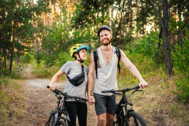 Genç beyaz bisikletçiler parktaki orman yolunda dağ bisikletlerini yürütür ve iterler. Aktif spor hafta sonu. Kırsalda bisikleti olan güzel insanlar..