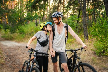 Genç beyaz bisikletçiler parktaki orman yolunda dağ bisikletlerini yürütür ve iterler. Aktif spor hafta sonu. Kırsalda bisikleti olan güzel insanlar..