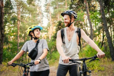 Bisiklet süren ve orman yolunda yürüyen neşeli bir çift. Bisikletli mutlu çift parkta yürüyor, konuşuyor ve gülüyor. Kafkasyalı genç adam ve kadın ormanda bisiklet sürüyor..