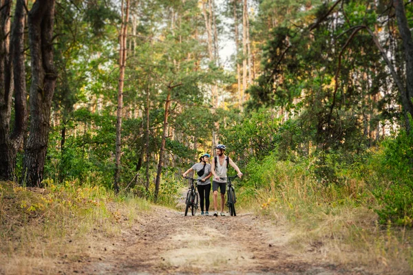 若い白人のカップルは 公園内の森林道路に沿ってマウンテンバイクを歩いてプッシュします スポーツの週末だ 田舎で自転車を持っているスポーティ美しい人々 — ストック写真