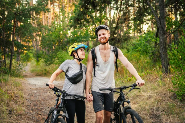 Genç beyaz bisikletçiler parktaki orman yolunda dağ bisikletlerini yürütür ve iterler. Aktif spor hafta sonu. Kırsalda bisikleti olan güzel insanlar..