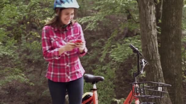 Una joven, hermosa mujer caucásica en ropa casual está descansando cerca de una bicicleta naranja de alquiler en el parque y utiliza un teléfono inteligente. Deportes y Tecnología. Mujer con bicicleta utilizando Internet en el teléfono inteligente — Vídeos de Stock