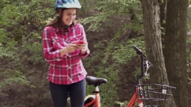 El ciclista usa el teléfono celular mientras monta en bicicleta. Mujer joven con bicicleta usando smartphone en el parque. Joven hembra en bicicleta en madera comprobando el teléfono celular. Una chica descansando en un bosque. Deportes y Tecnología — Vídeo de stock