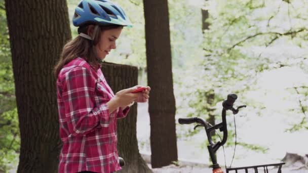 뜻하지 않은 옷을 입은 젊고 아름다운 코카서스 여성이 공원에 있는 한빌린 오렌지 자전거 근처에서 휴식을 취하고 있으며 스마트폰을 사용하고 있다. 스포츠와 기술. 자전거를 타고 스마트폰으로 인터넷을 하는 여성 — 비디오