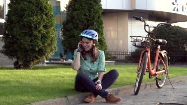 İnsan ve şehir bisikleti, çevre dostu ulaşım. Çimlerin üzerinde dinlenen güzel beyaz kadın işçi turuncu bisikletli kırmızı bir cep telefonu kullanıyor.