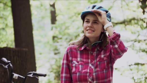 Ung kvinna med cykel står i parken. Porträtt av ung vacker flicka står nära cykel, poserar och ler, bär en röd rutig skjorta i skogen. Ämne ekologiskt transportsätt cykel — Stockvideo