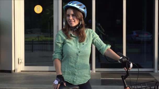 Temat do pracy na rowerze. Młoda kaukaska kobieta przyjechała do biura na ekologicznym rowerze transportowym. Dziewczyna w biurowcu parkingu dla rowerów w kasku, rękawiczkach, koszuli i dżinsach — Wideo stockowe