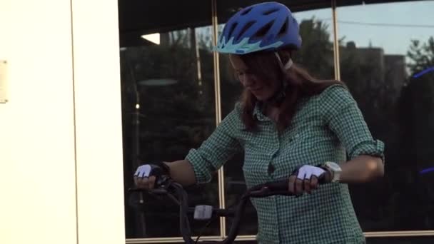 Tema para trabajar en la moto. Una joven mujer caucásica llegó en bicicleta de transporte respetuosa con el medio ambiente a la oficina. Chica en un edificio de oficinas de estacionamiento de bicicletas en un casco, guantes y camisa y jeans — Vídeo de stock