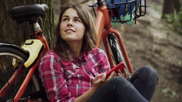 Transporte ecológico en bicicleta. Joven estudiante caucásica sentarse descansando en el parque cerca de alquiler bicicleta naranja, el uso de teléfono móvil. Dama sonriente irrumpiendo en el césped en el bosque. Vacaciones, viajes, redes sociales — Vídeo de stock