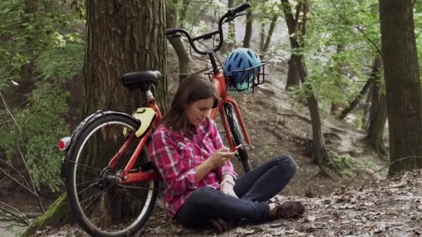 Экологический велосипедный транспорт. Молодая кавказская студентка сидит в парке рядом с арендой оранжевого велосипеда, пользуется мобильным телефоном. Улыбающаяся леди, лежащая на лужайке в лесу. Отдых, путешествия, социальные сети — стоковое видео