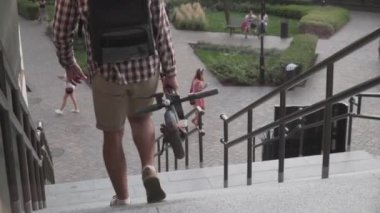 Modern binanın yakınlarında katlanmış elektrikli scooterla yürüyen bir adam. Ekolojik teknolojik yaşam tarzı. E-Hareket kabiliyeti. Elektrikli Scooter 'ı taşıyan kişi katlanmış pozisyonda. Çevre taşımacılığı