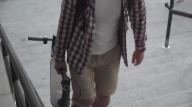 Katlanmış bir E-scooter taşıyan bir adam binanın yanındaki merdivenlerde. Çevresel şehir taşımacılığı, modern şehir konsepti. Ekolojik teknolojik yaşam tarzı. Katlanmış Konumda Elektrikli Scooter Taşıyıcı
