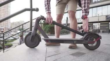Modern binanın yakınlarında katlanmış elektrikli scooterla yürüyen bir adam. Ekolojik teknolojik yaşam tarzı. E-Hareket kabiliyeti. Elektrikli Scooter 'ı taşıyan kişi katlanmış pozisyonda. Çevre taşımacılığı
