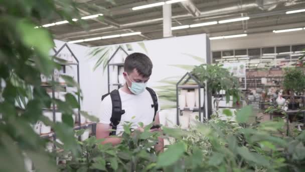 白人男性の購入者は、コヴィド19パンデミックの際に顔の盾に保育園の観葉植物を選択する。コロナウイルスの流行や感染を保護するために呼吸器を身に着けているガーデンセンターでの男のショッピング工場 — ストック動画