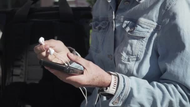 Ώριμη γυναίκα ξοδεύει τον ελεύθερο χρόνο της ακούγοντας ενεργά μουσική με ακουστικά και οδηγώντας ένα ηλεκτρικό σκούτερ στην πόλη. Μεσήλικες γυναίκες κάθονται στον πάγκο κρατώντας smartphone στα χέρια και χρησιμοποιεί αεροθαλάμους — Αρχείο Βίντεο
