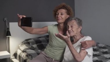 Mutlu anne ve kız COVID 19 sırasında akıllı telefon kullanıyor. Neşeli orta yaşlı ve yaşlı kadınlar evde cep telefonuyla görüntülü konuşma sırasında el sallıyorlar. Yaşlı bayana cep telefonu kullanmayı öğreten kadın.