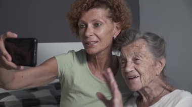 Mutlu anne ve kız COVID 19 sırasında akıllı telefon kullanıyor. Neşeli orta yaşlı ve yaşlı kadınlar evde cep telefonuyla görüntülü konuşma sırasında el sallıyorlar. Yaşlı bayana cep telefonu kullanmayı öğreten kadın.