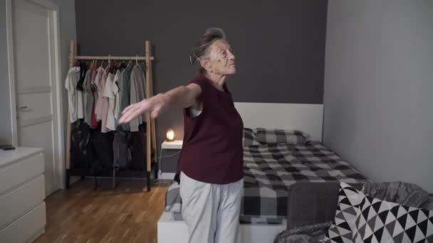 Дуже стара жінка з сірим волоссям і глибокими зморшками робить фізичні вправи вдома. Літня жінка розігрівається в маленькій квартирі. Щасливий старший тренування фітнес в дверях під час ковадла 19 — стокове відео