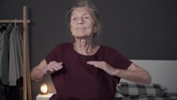 白髪と深いしわを持つ非常に古い年齢の女性は自宅で物理的な演習を行います。年配の女性が小さなアパートで暖まっている。covid 19中のドア内のハッピーシニア運動フィットネス — ストック動画