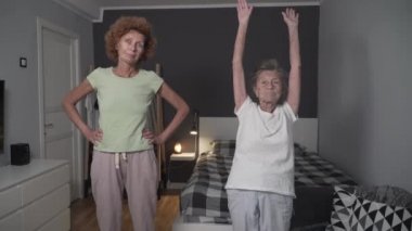 Yaşlı insanlar için egzersiz. İki kadın, olgun kız ve emekli anne, evdeki küçük odada sağlık egzersizleri yapıyorlar. Yaşlı, kır saçlı kadın bakımevinde hemşireyle sağlıklı bir yaşam sürüyor.