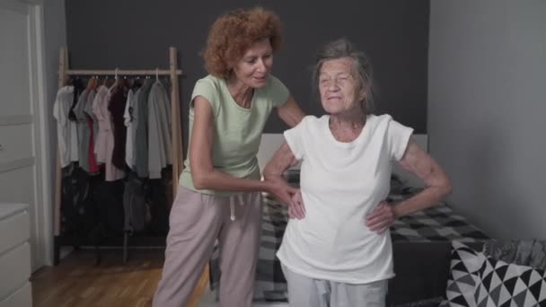 老人のための運動。2人の女性、成熟した娘と引退した母親は、自宅の小さな部屋で健康のための演習を行います。老人ホームで看護師と健康的な生活を実践シニア白髪の女性 — ストック動画