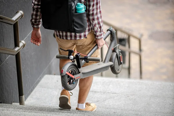 Man walking with folded electric scooter up stairs in city near modern building. Ecological technological lifestyle. E-Mobility. Person Carrying Electric Scooter Folded Position. Eco urban transport.